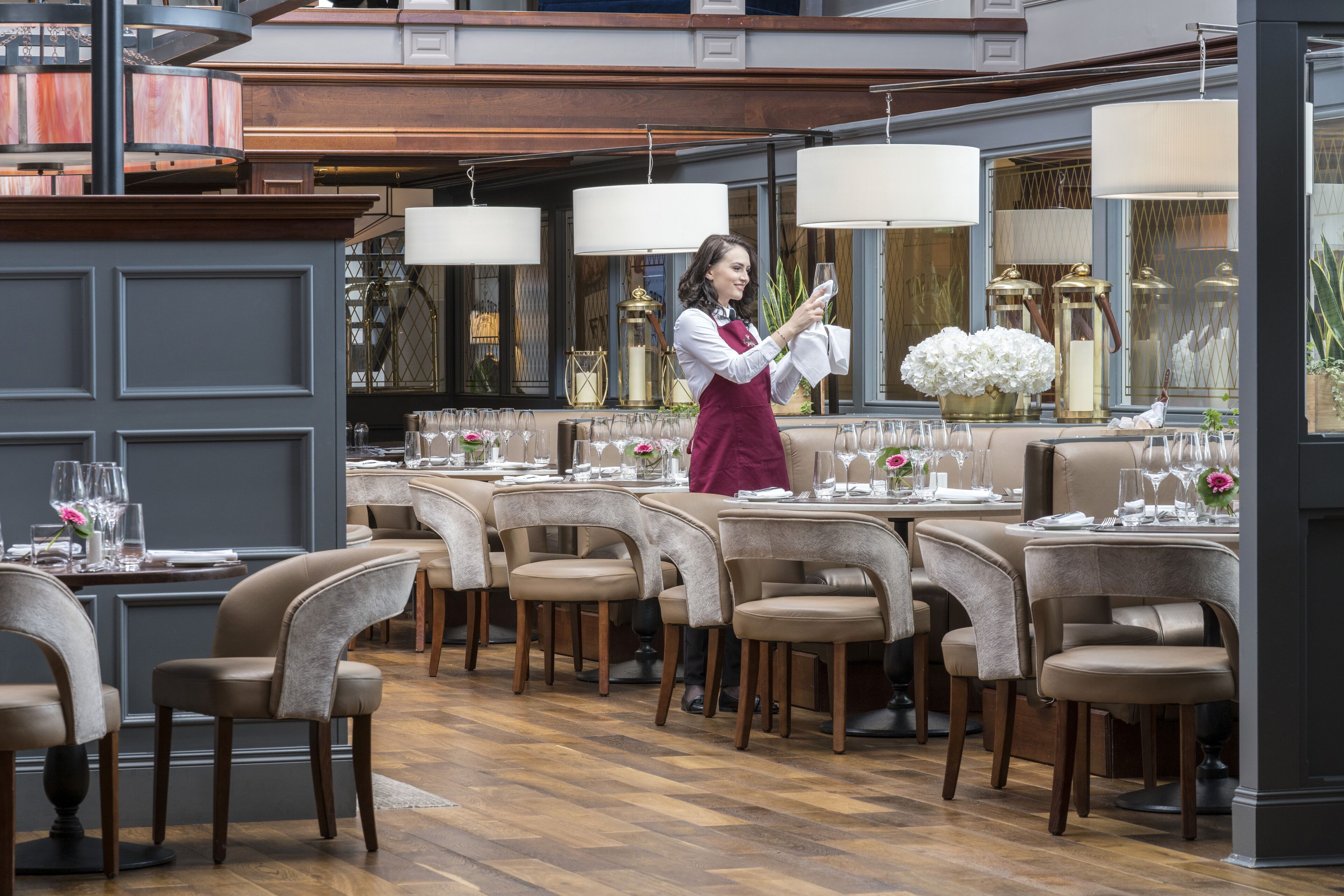 Un restaurant avec de nombreuses tables dressées pour le service, un serveur portant un tablier rouge et un décor intérieur élégant.