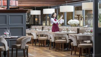 Un restaurant avec de nombreuses tables dressées pour le service, un serveur portant un tablier rouge et un décor intérieur élégant.
