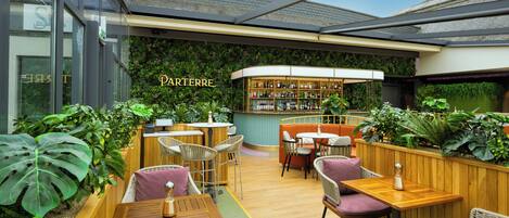 Un rincón al aire libre acogedor con mesas y sillas de madera, rodeado de abundante vegetación y una zona de bar con un letrero que dice 'PARTERRE'.