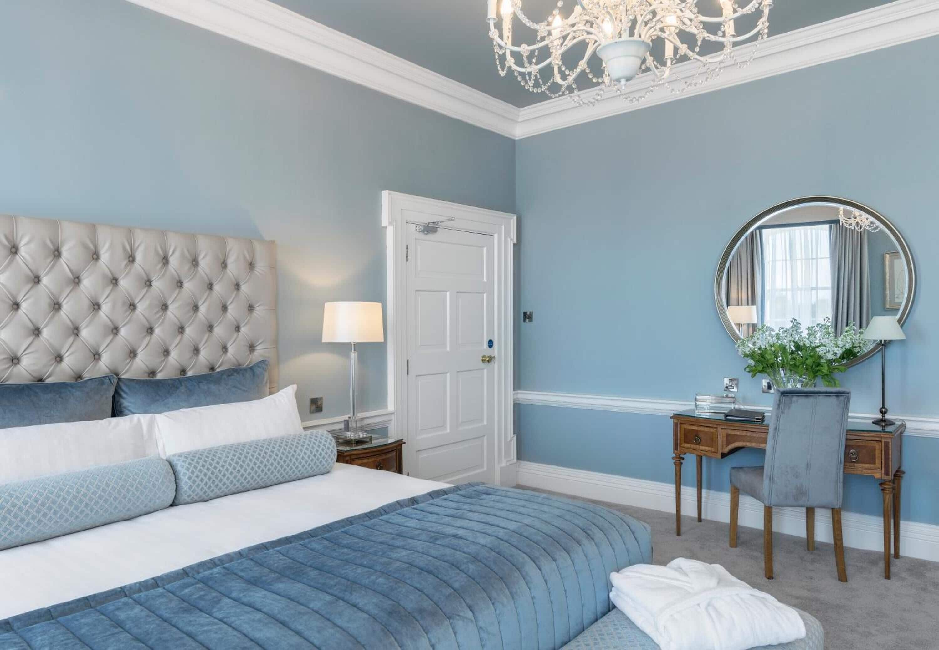 A bedroom with a tufted headboard, a large bed with blue bedding, a desk with a chair, a round mirror, and a chandelier.