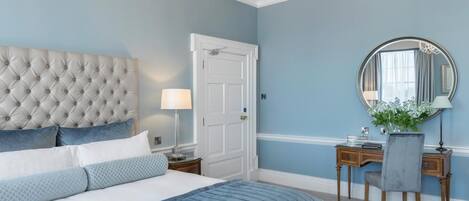 A bedroom with a tufted headboard, a large bed with blue bedding, a desk with a chair, a round mirror, and a chandelier.