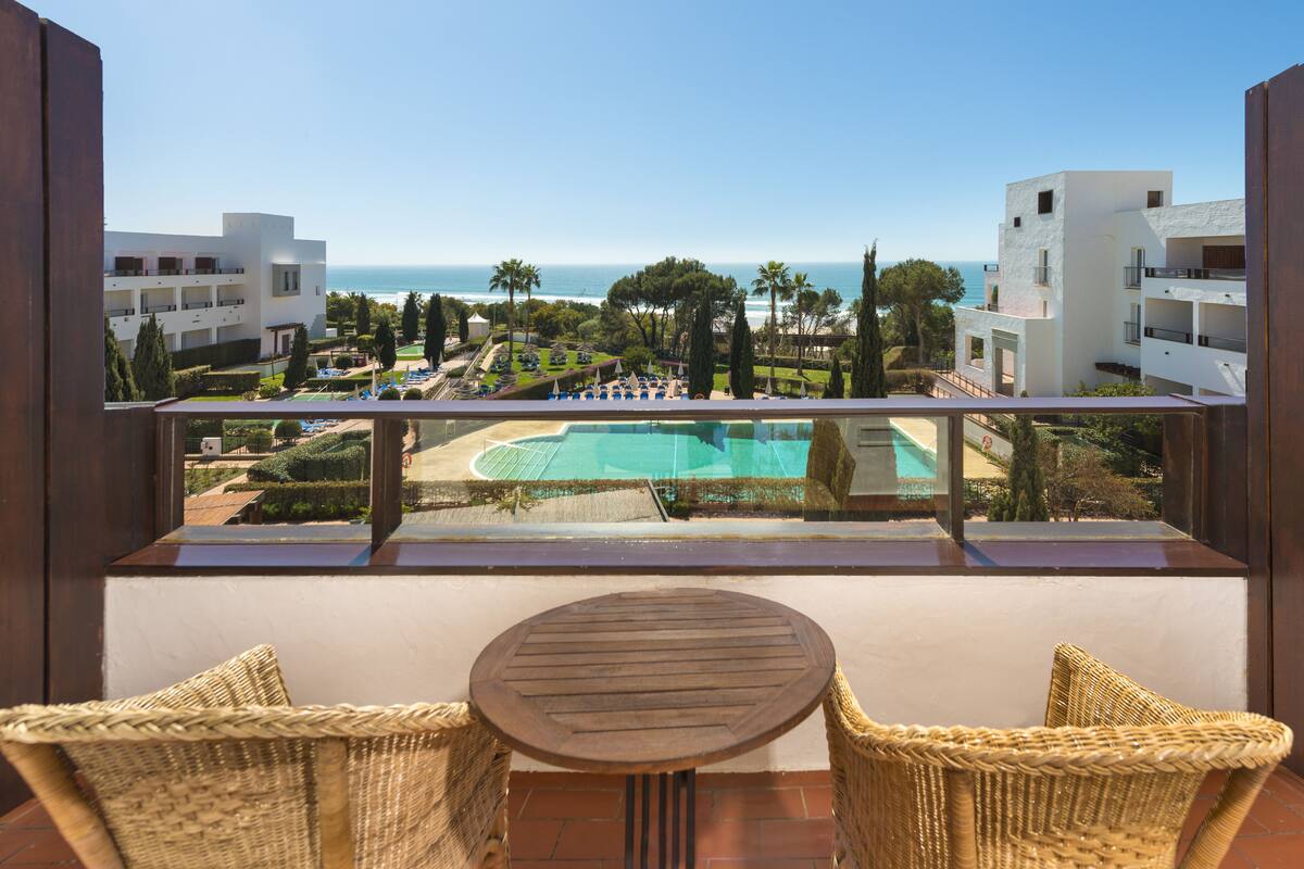 A balcony with wicker chairs and a small table, overlooking a pool and a landscaped area with trees and a view of the sea.
