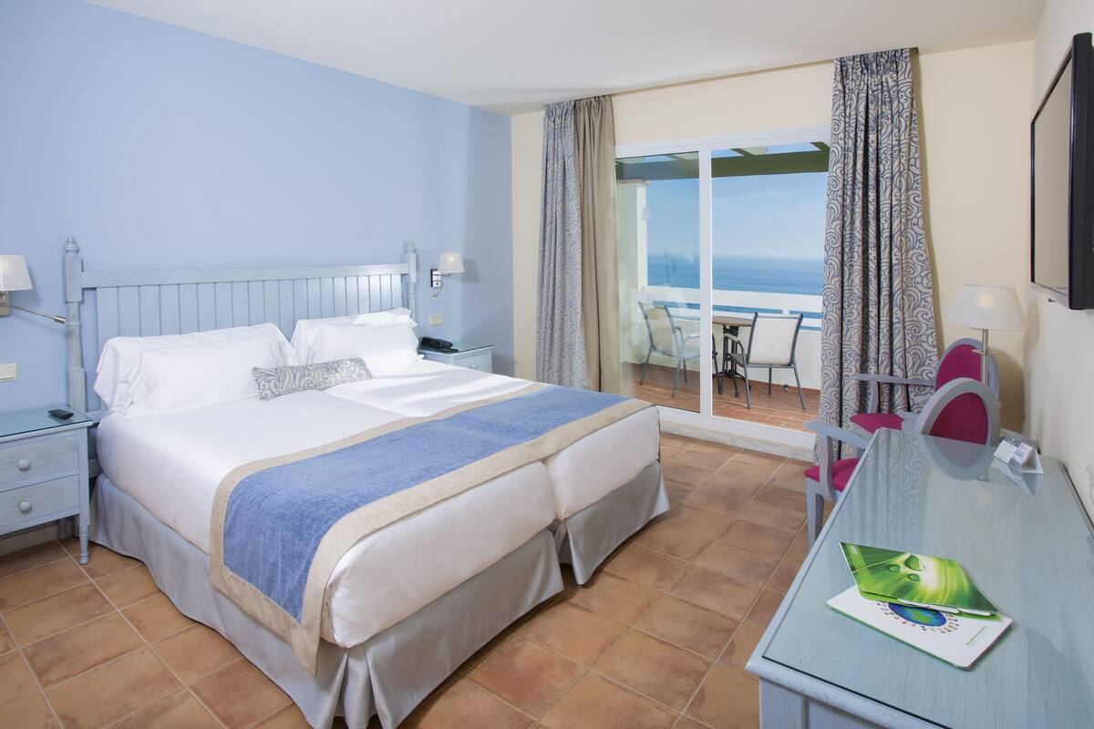 A hotel room with a bed, a desk with a chair, a window with curtains, and a view of the sea.