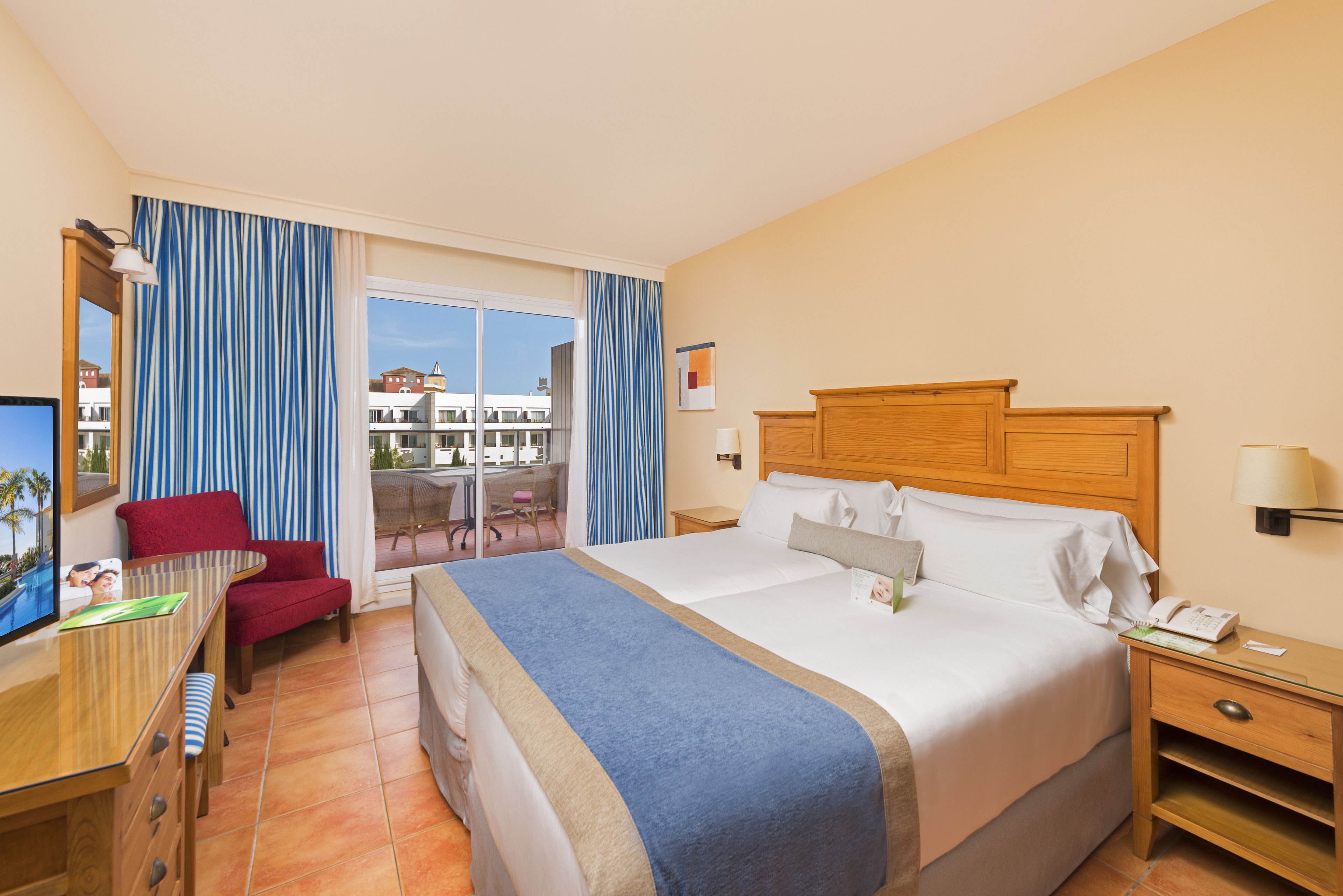 Habitación de hotel con una cama grande, un escritorio con computadora, una silla roja y una ventana con cortinas.