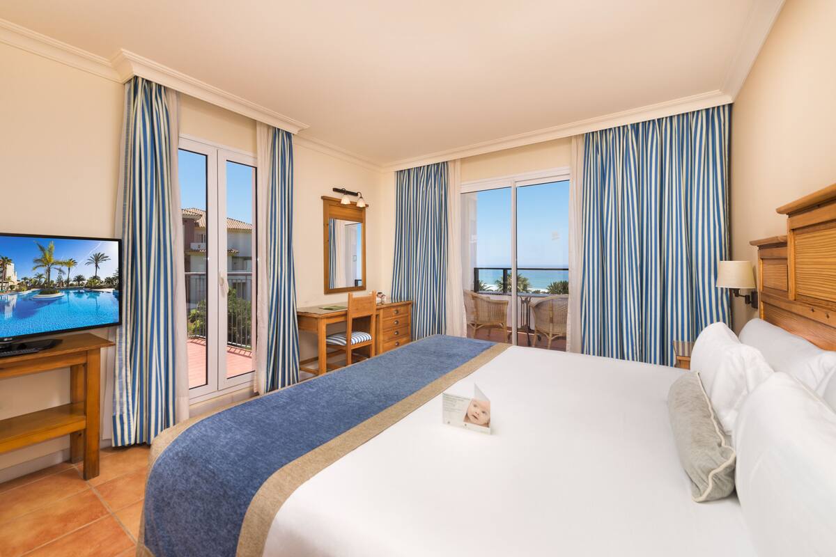 A hotel room with a large bed, a desk, a television, and a balcony with a view of the ocean.