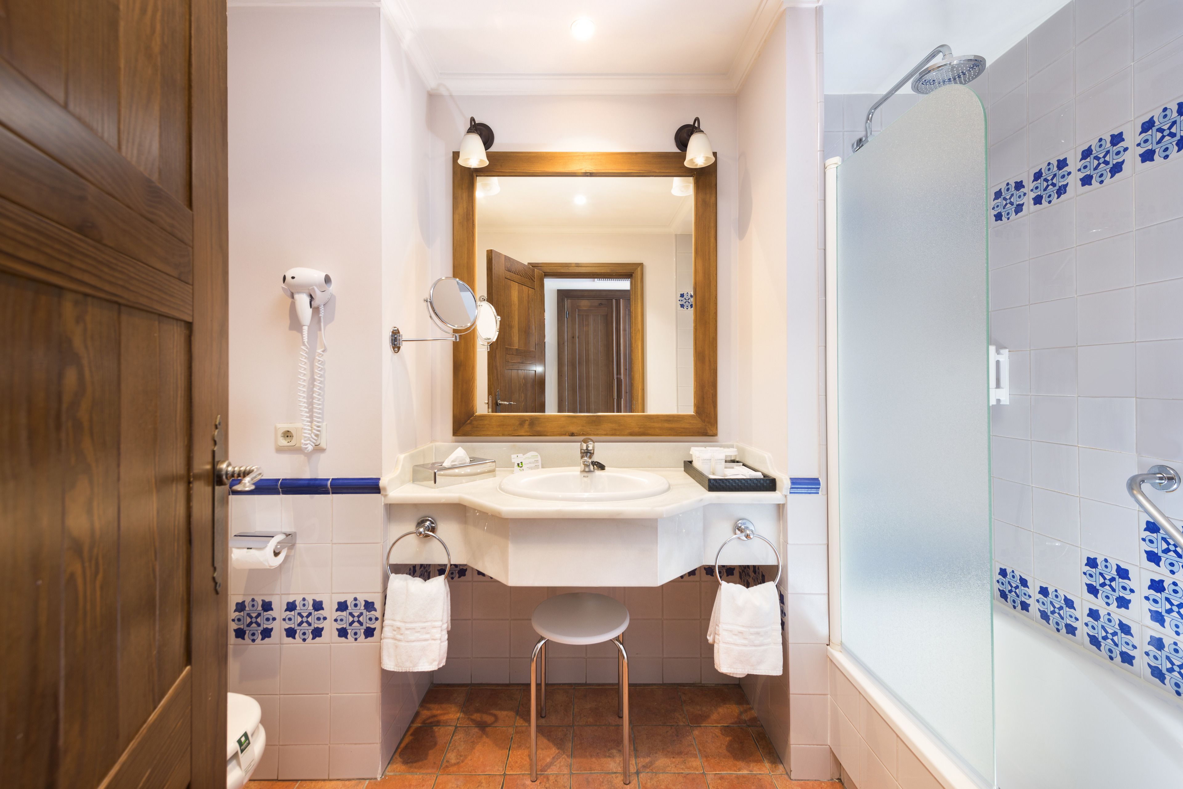 A bathroom with a wooden door, a white sink with a mirror above it, a glass shower enclosure, and a small stool.