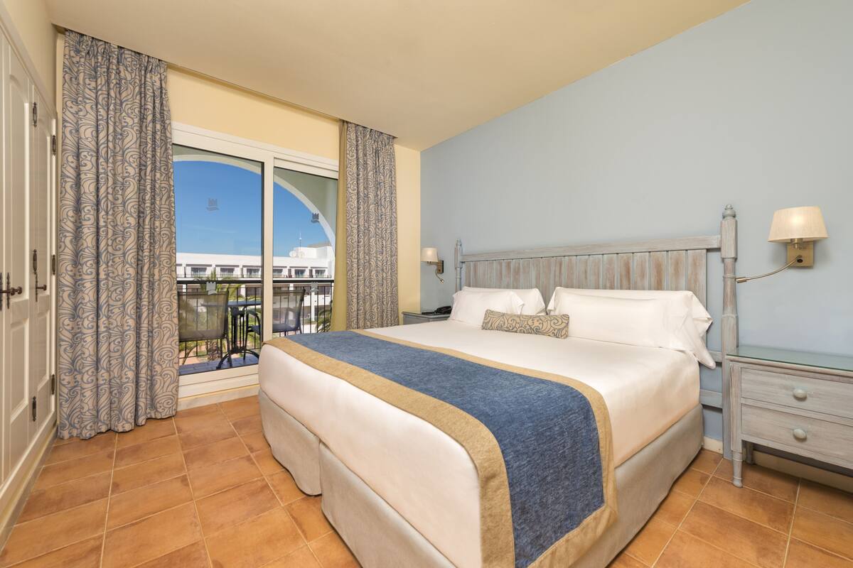 A hotel room with a large bed, a nightstand, a lamp, and a view of the outside.