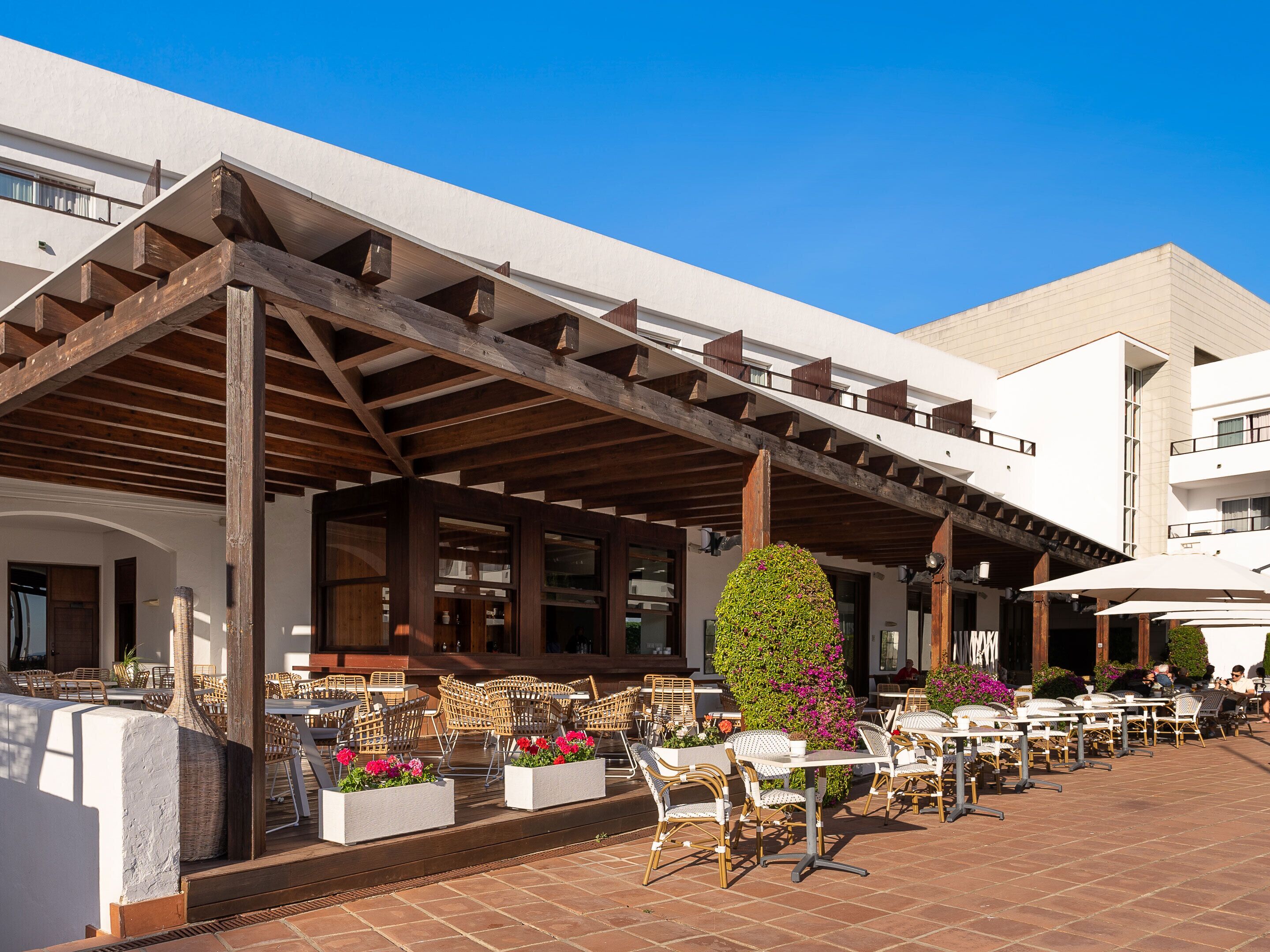 An outdoor seating area with wicker chairs and tables, a covered patio with wooden beams, and a building with white walls and balconies.