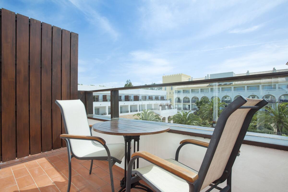 A balcony with two chairs and a table, overlooking a courtyard with buildings and palm trees.
