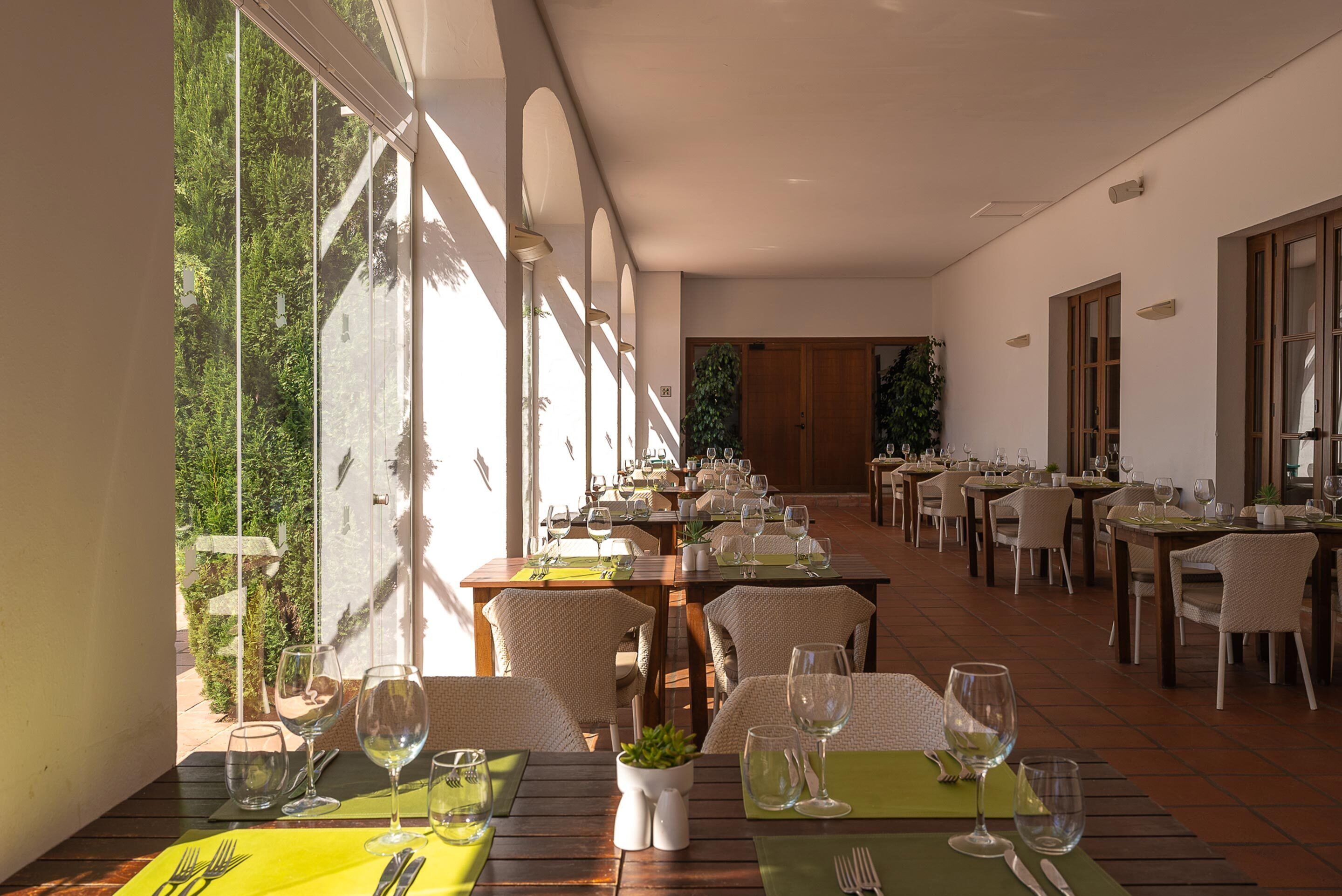 A restaurant with multiple dining tables, chairs, and a view of greenery through large windows.