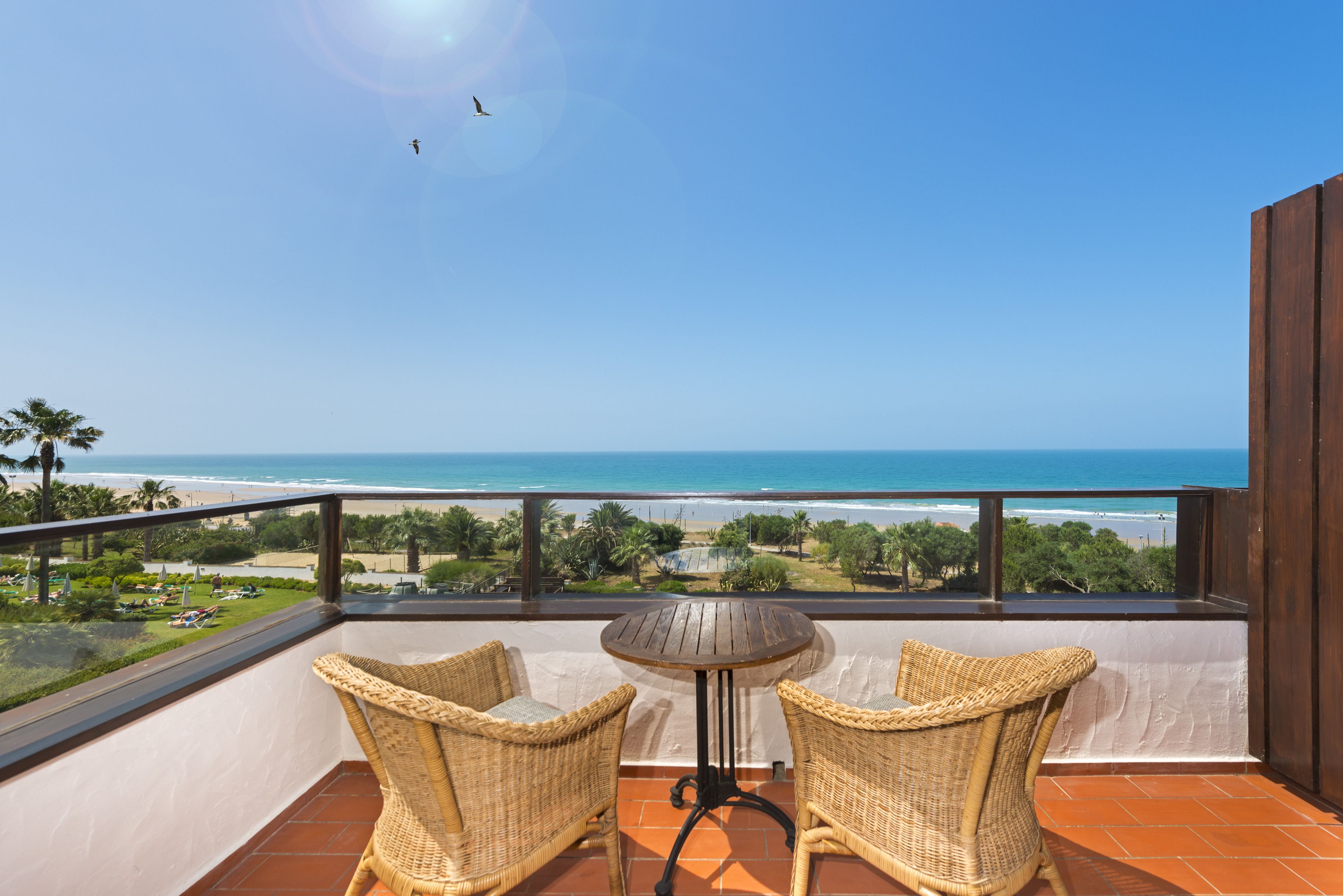 A balcony with wicker chairs and a small table, overlooking a beach and ocean.