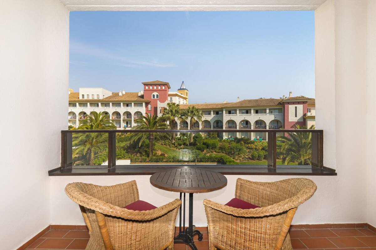 A balcony with wicker chairs and a table, overlooking a courtyard with buildings and palm trees.