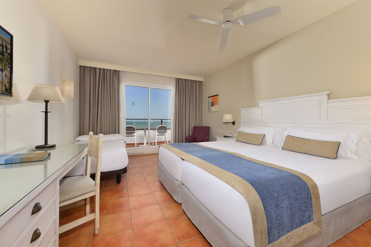 A hotel room with a bed, desk, chair, and a view of the sea through the window.