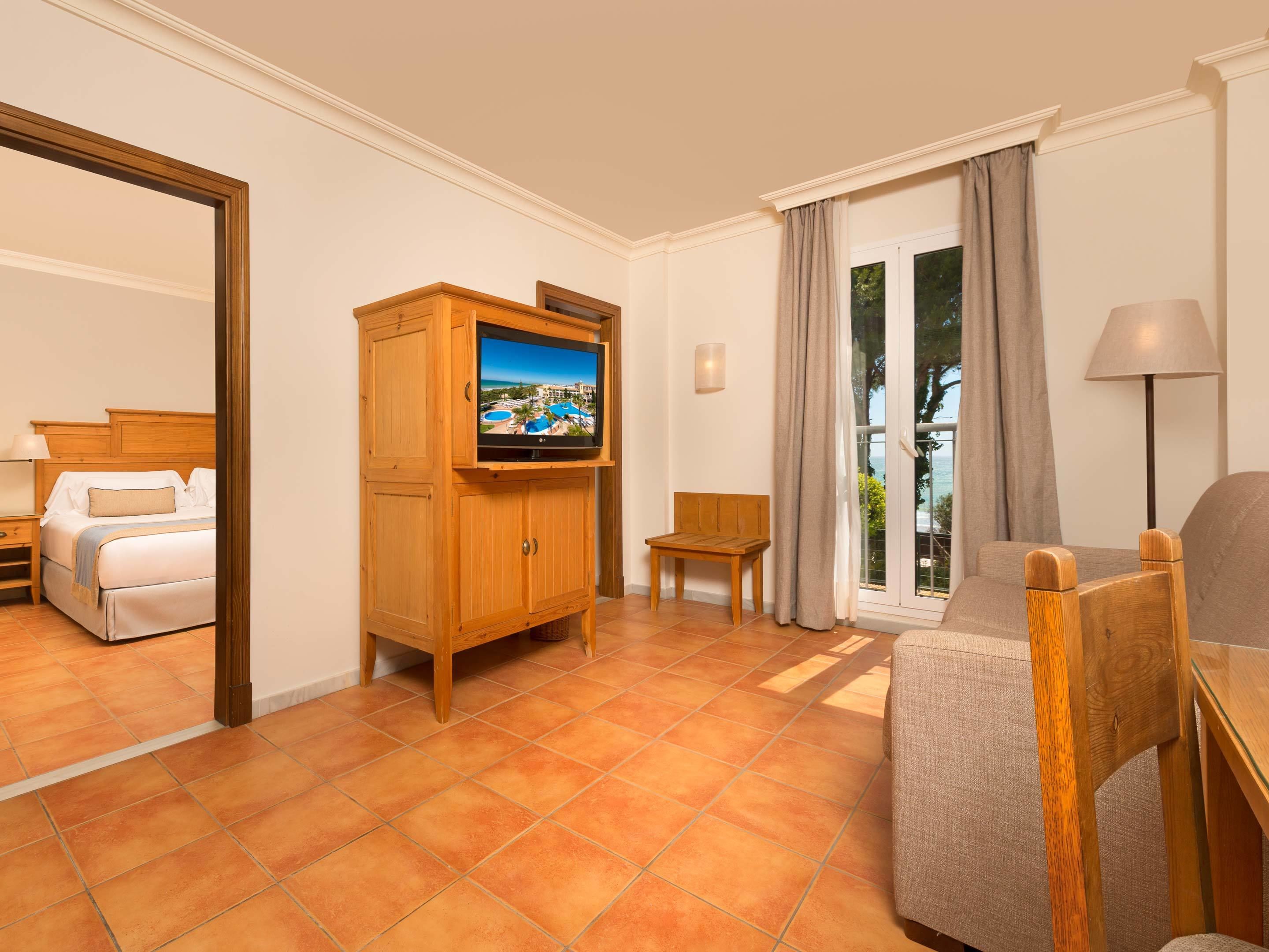 A hotel room with a bed, a TV on a wooden cabinet, a chair, a sofa, and a view of the outdoors through a glass door.