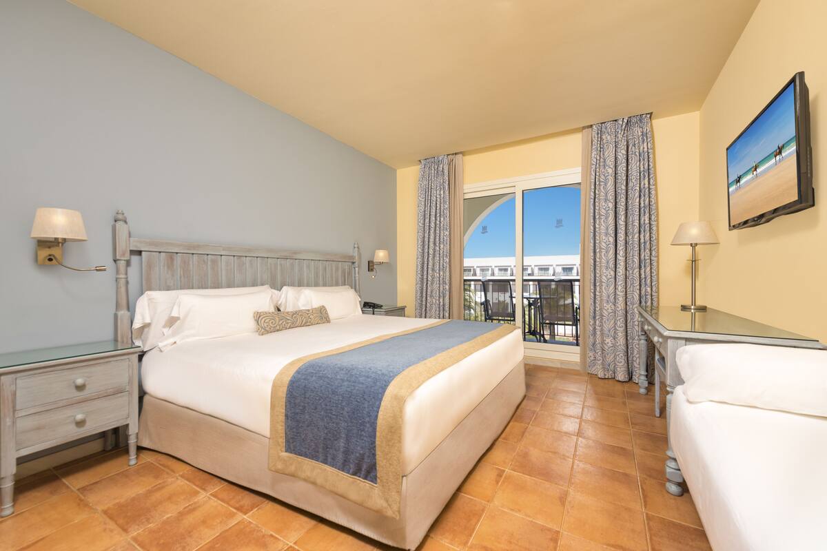 A hotel room with a large bed, a desk, a chair, and a balcony with a view of the beach.