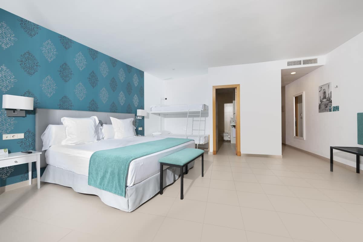 A hotel room with a bed, bedside table, bench, and a bathroom visible through an open door.