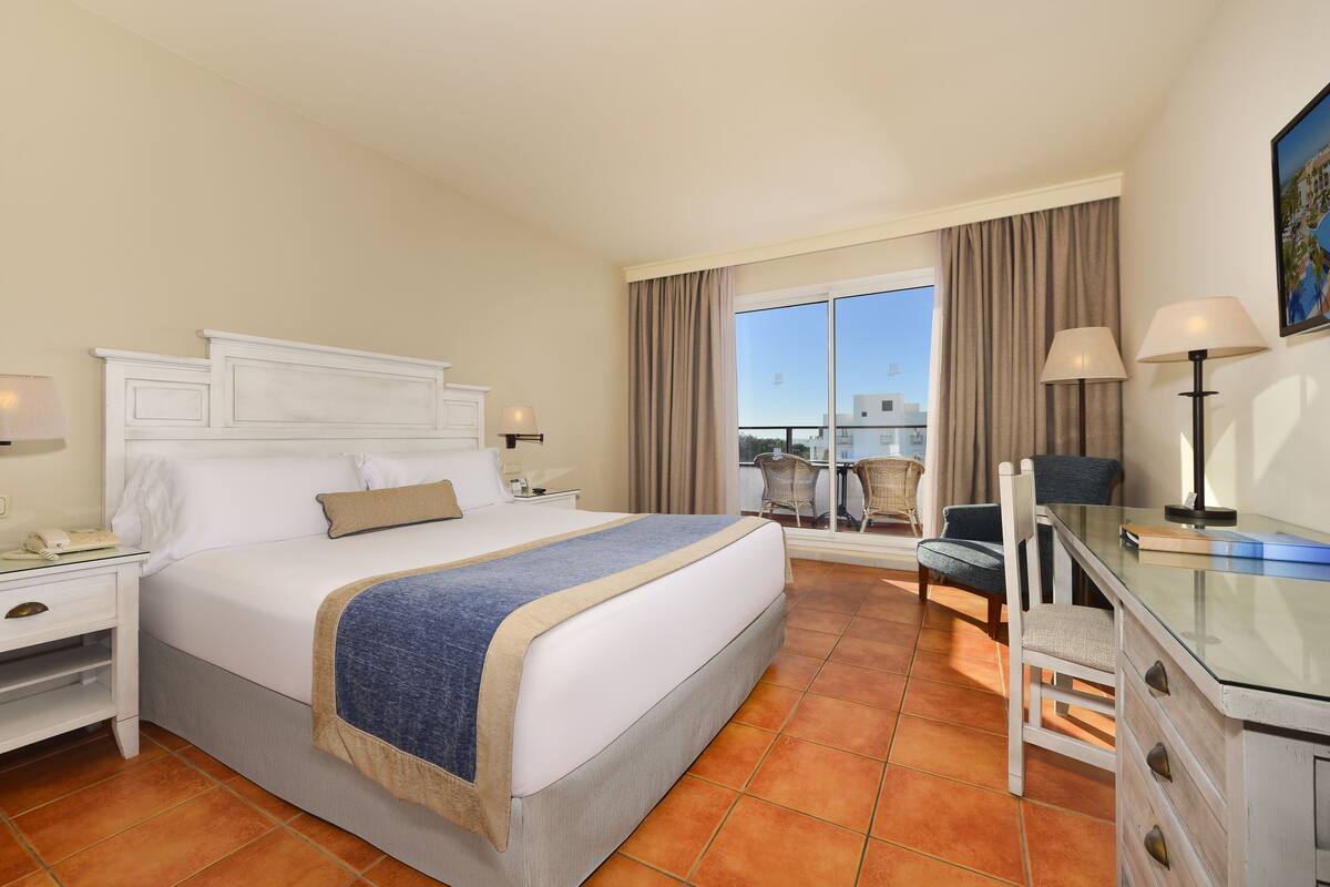 A hotel room with a bed, a desk, a chair, a nightstand, and a view of the outside.