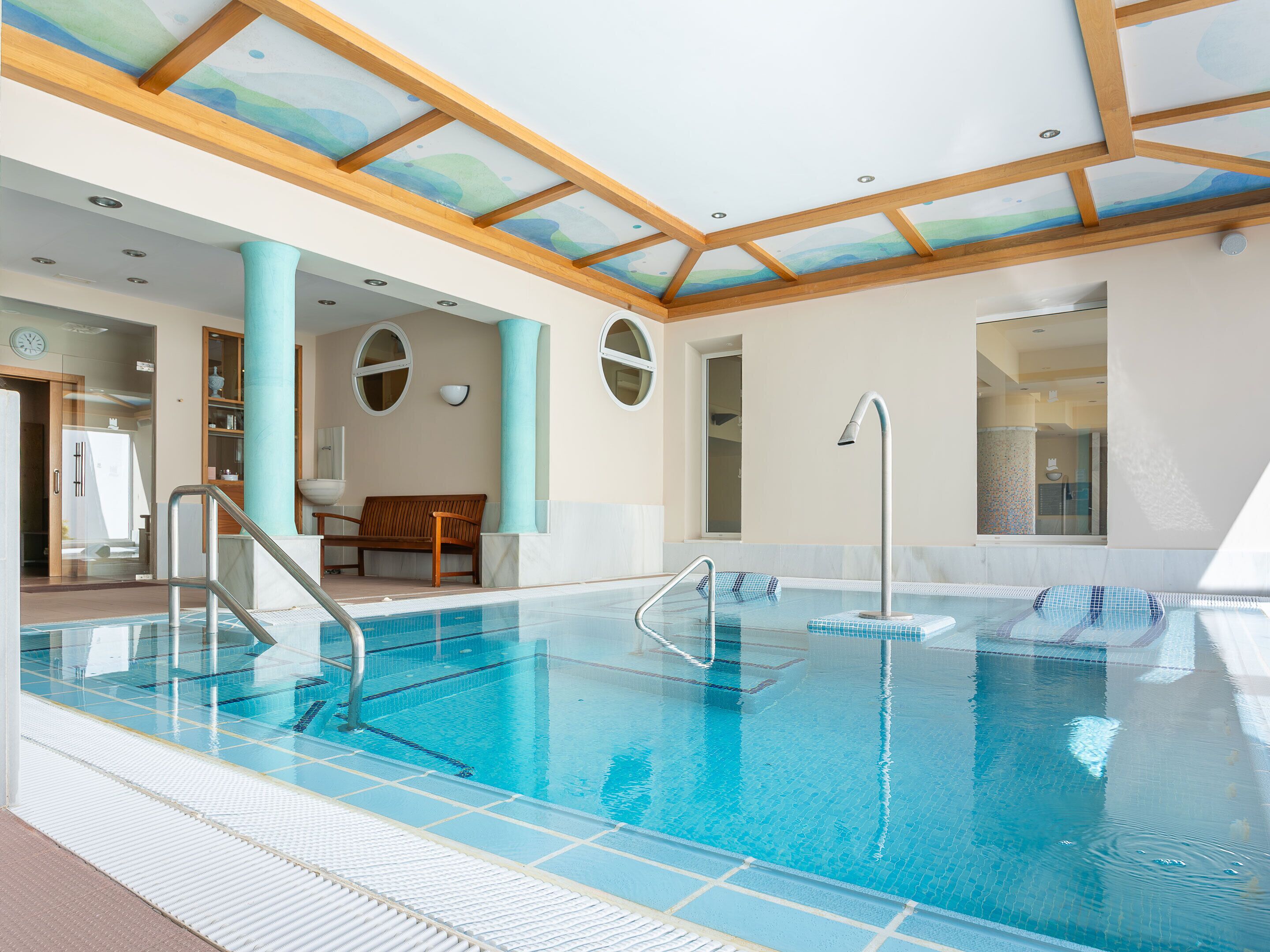 A large indoor swimming pool with clear blue water, wooden beams on the ceiling, and a bench near the entrance.