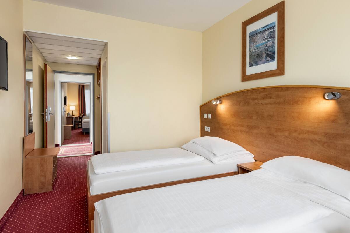 Habitación de hotel con dos camas individuales, cabecera de madera, suelo cubierto de moqueta roja, pasillo que conduce a un baño y un cuadro enmarcado en la pared.