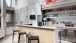 Eine moderne Bar mit einer Holztheke, zwei schwarzen Barhocker, einem an der Wand befestigten Fernseher und einer Auswahl an Flaschen und Gläsern auf Regalen.