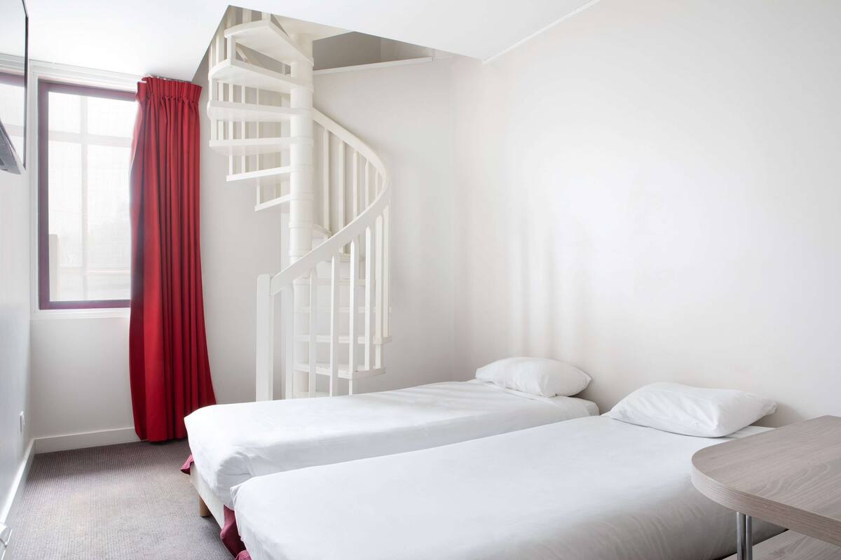 Habitación de hotel con escalera espiral blanca, dos camas con ropa de cama blanca, una cortina roja y una mesa de madera.
