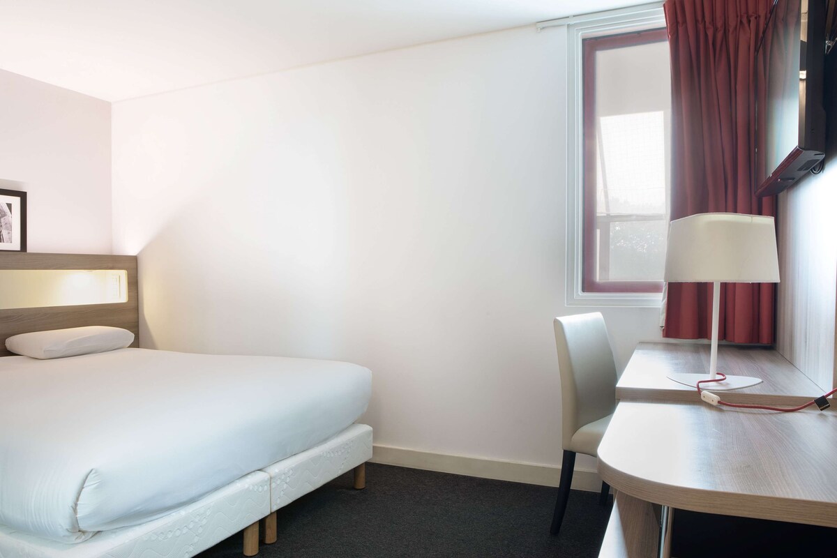 Habitació d'hotel amb llit, escriptori, cadira, làmpada i finestra amb cortines.