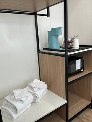 Habitación de hotel con kitchenette, microondas, hervidor de agua y una pila de toallas sobre un banco.