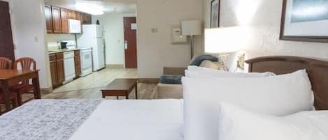 A hotel room with a bed, a desk, a chair, a sofa, and a kitchenette in the background.