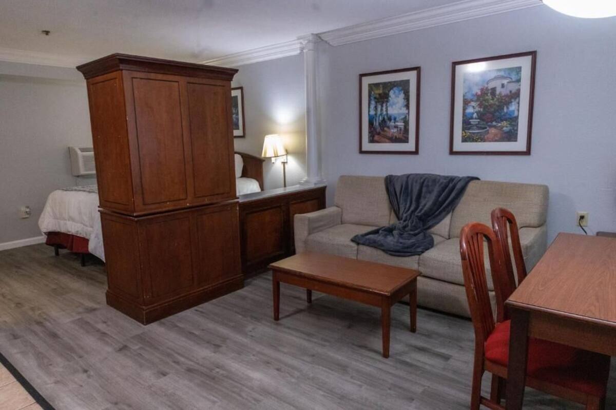 Ein Wohnzimmer mit einem Holzschrank, einer Couch, einem Couchtisch und einem Esstisch mit Stühlen. An der Wand hängen zwei gerahmte Bilder.