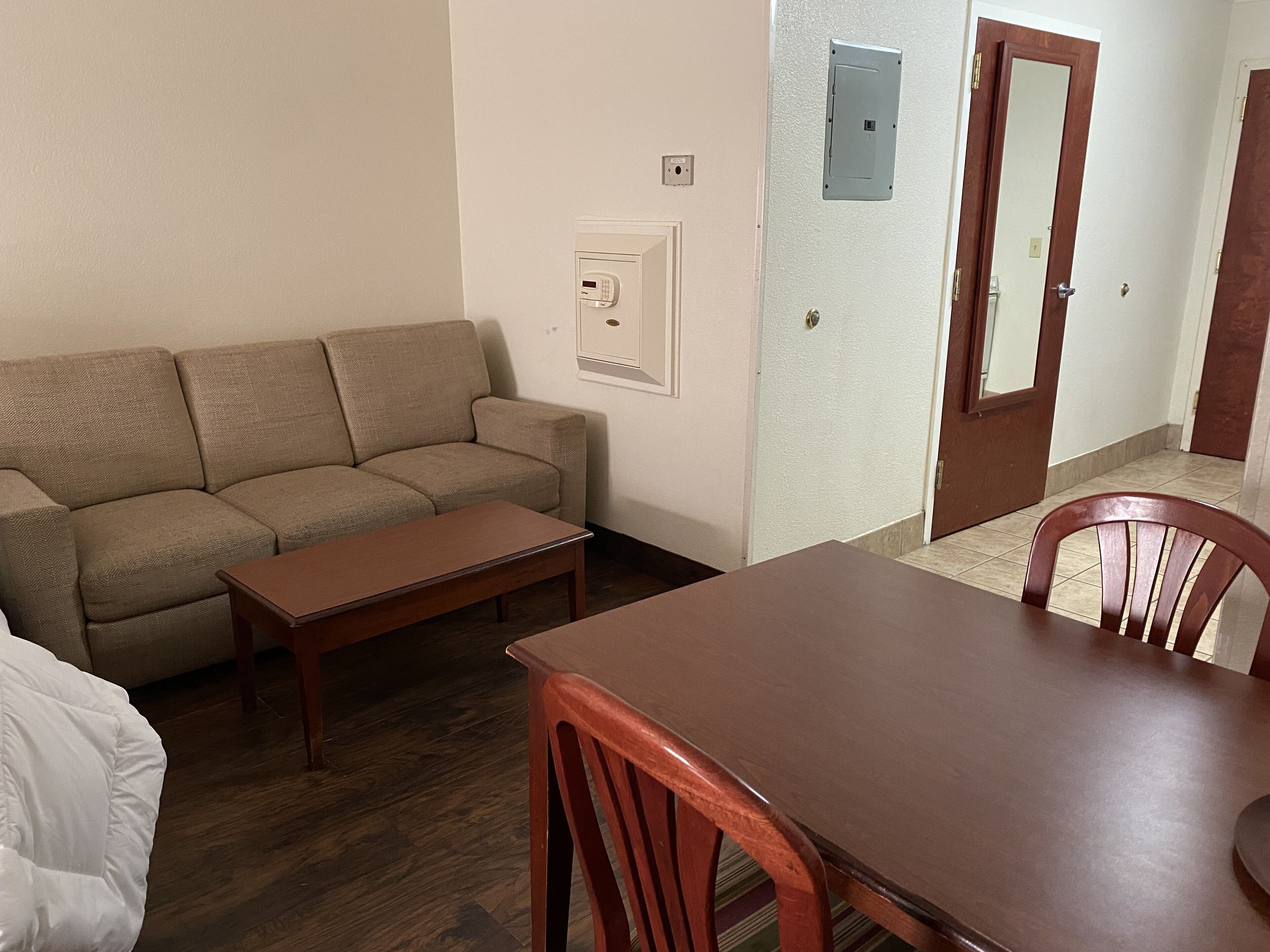 A living room with a sofa, a wooden table, and a chair. There is a door with a mirror and a light switch on the wall.