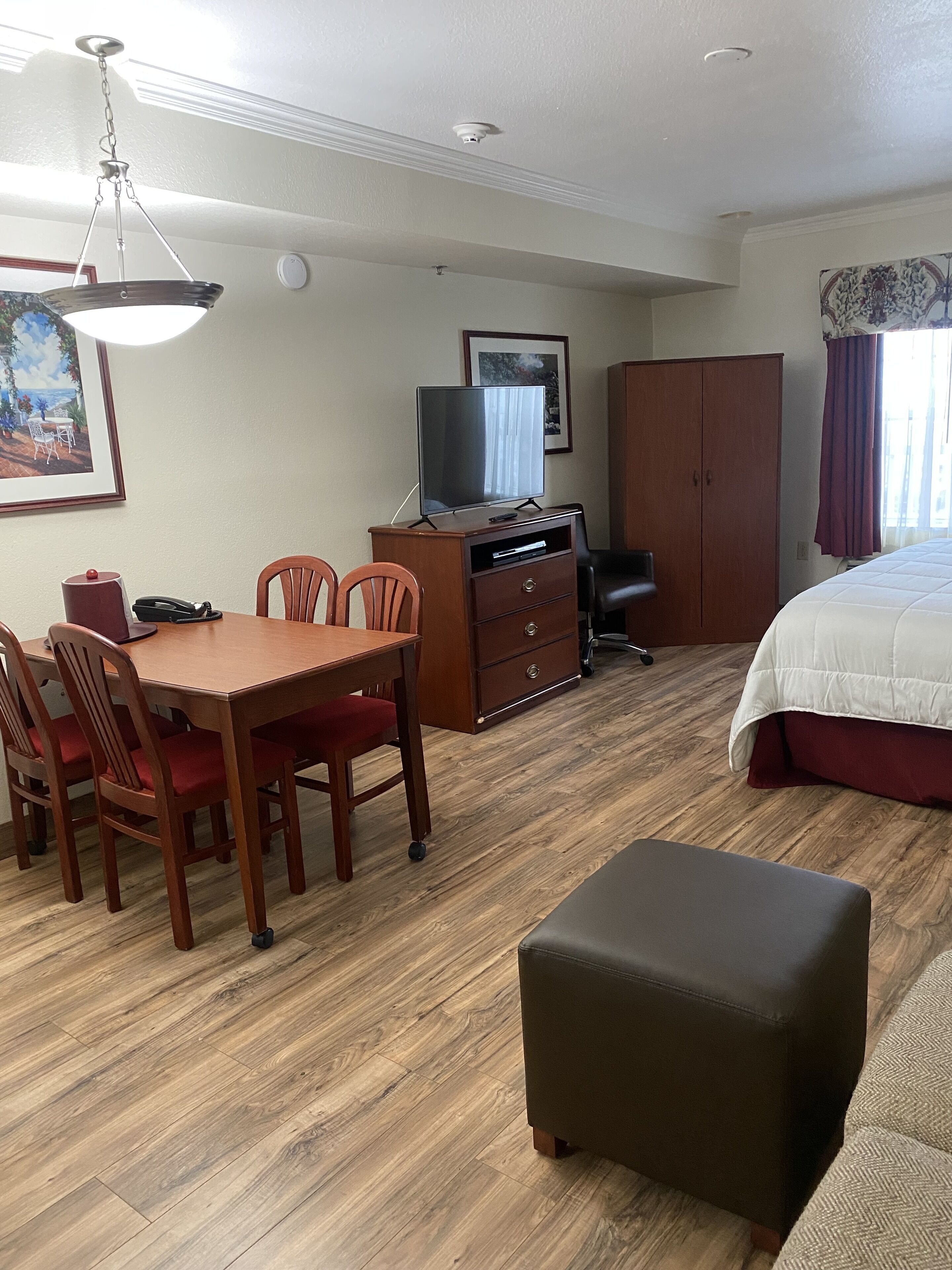 A hotel room with a wooden dining table, chairs, a TV on a wooden cabinet, a wardrobe, a bed with a red bedspread, and a wooden floor.
