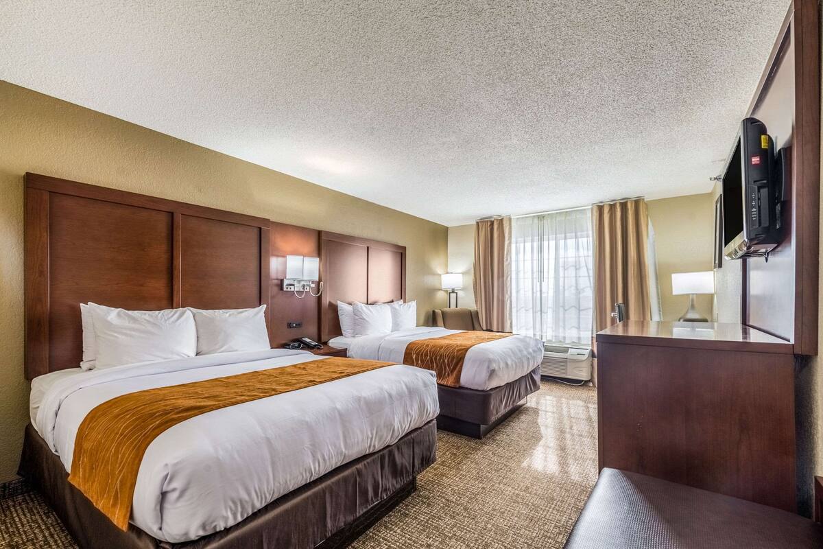 Une chambre d’hôtel avec deux lits, une tête de lit en bois, un téléviseur à écran plat, un bureau et une grande fenêtre avec des rideaux.