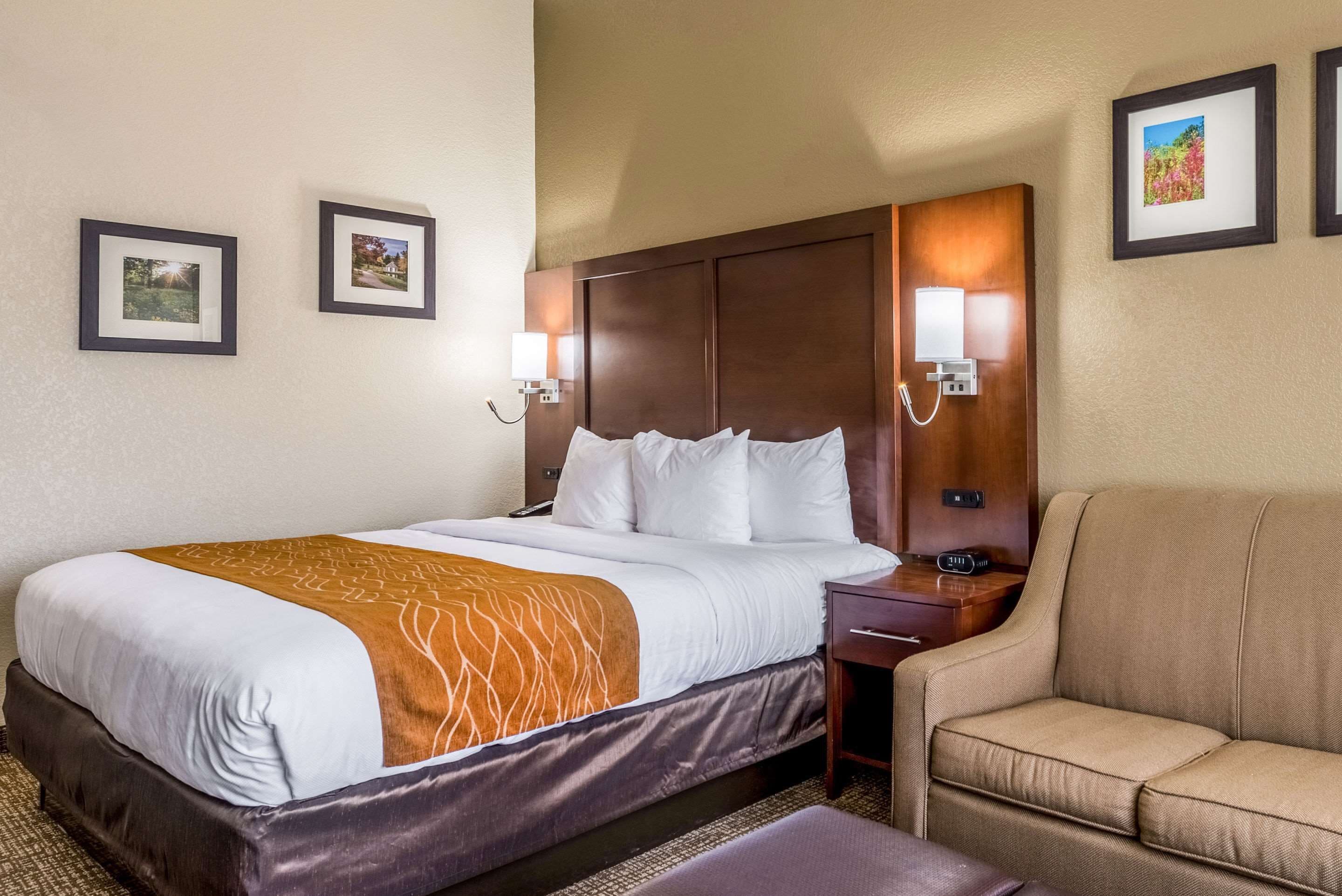 A hotel room with a large bed, a wooden headboard, two wall-mounted lamps, a small desk, and a sofa.