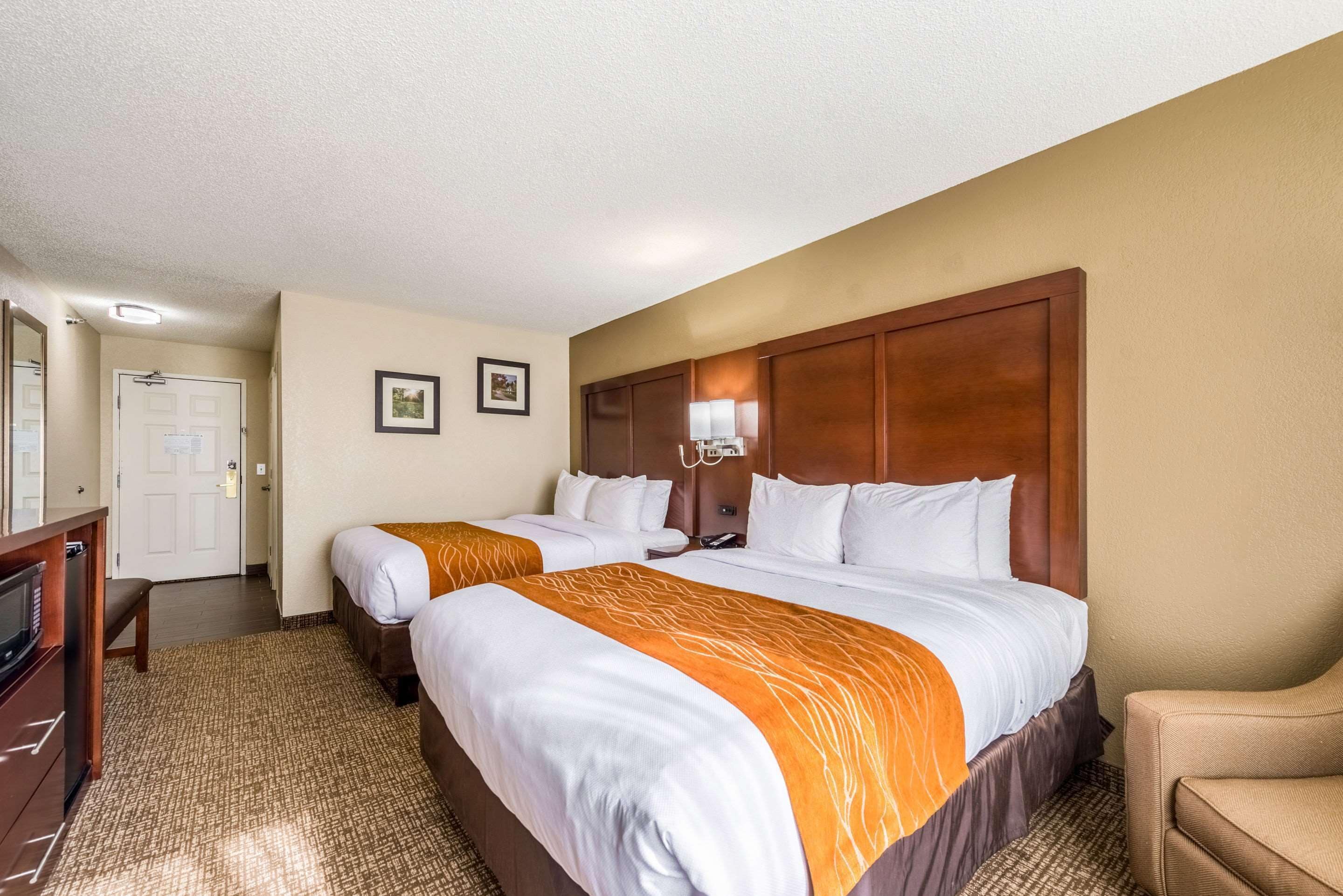 Quarto de hotel com duas camas, um painel de cabeceira de madeira, uma escrivaninha com abajur e uma cadeira.