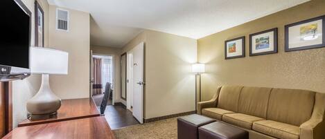 Une chambre d’hôtel avec un bureau en bois, un canapé beige, un téléviseur à écran plat et des tableaux encadrés au mur.