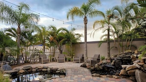 A patio area with a brick pathway, a small pond, lounge chairs, and palm trees.