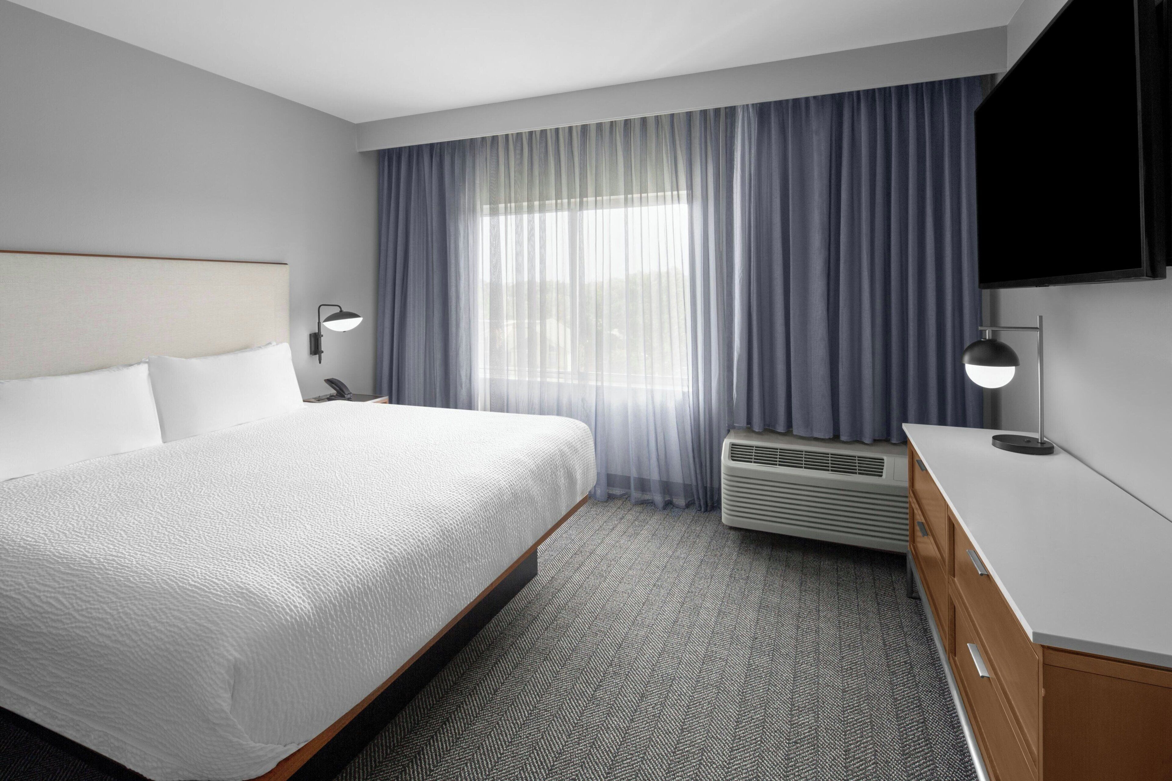 Ein Hotelzimmer mit einem großen Bett, einem Flachbildfernseher, einem Schreibtisch und einem Fenster mit Vorhängen.