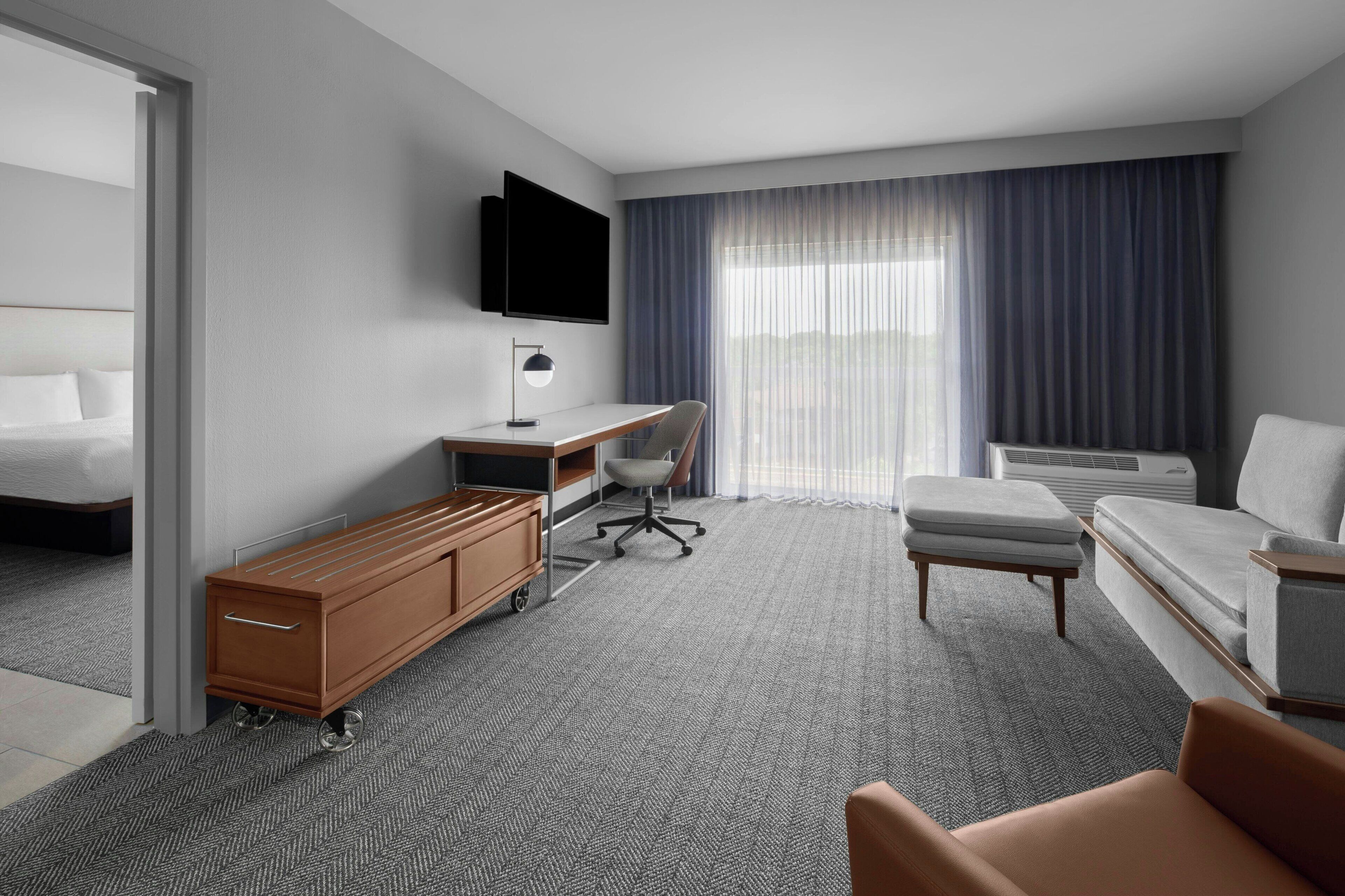 Een hotelkamer met een bed, een bureau met stoel, een bank, een aan de muur bevestigde televisie en een groot raam met gordijnen.