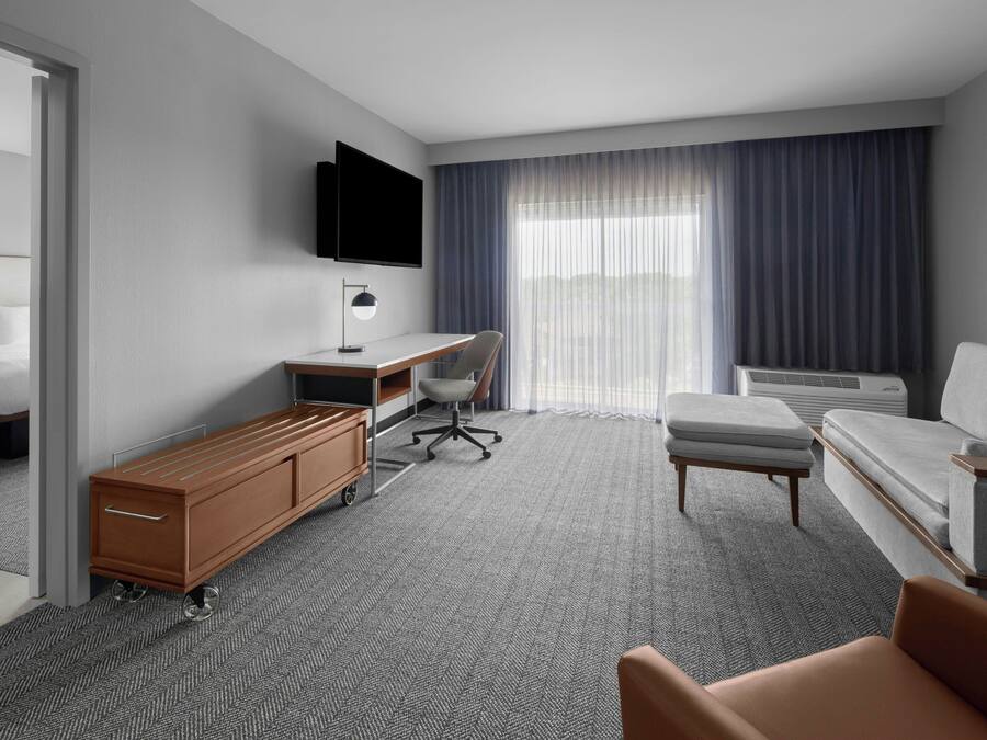 Een hotelkamer met een bed, een bureau met stoel, een bank, een aan de muur bevestigde televisie en een groot raam met gordijnen.