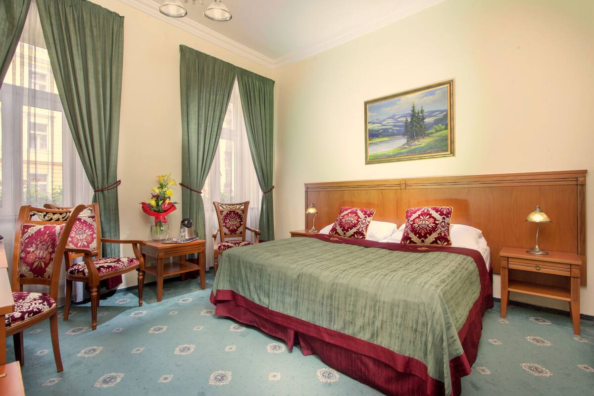 Habitación de hotel con una cama grande, dos mesitas de noche, un cabecero de madera, un cuadro en la pared y una ventana con cortinas verdes.