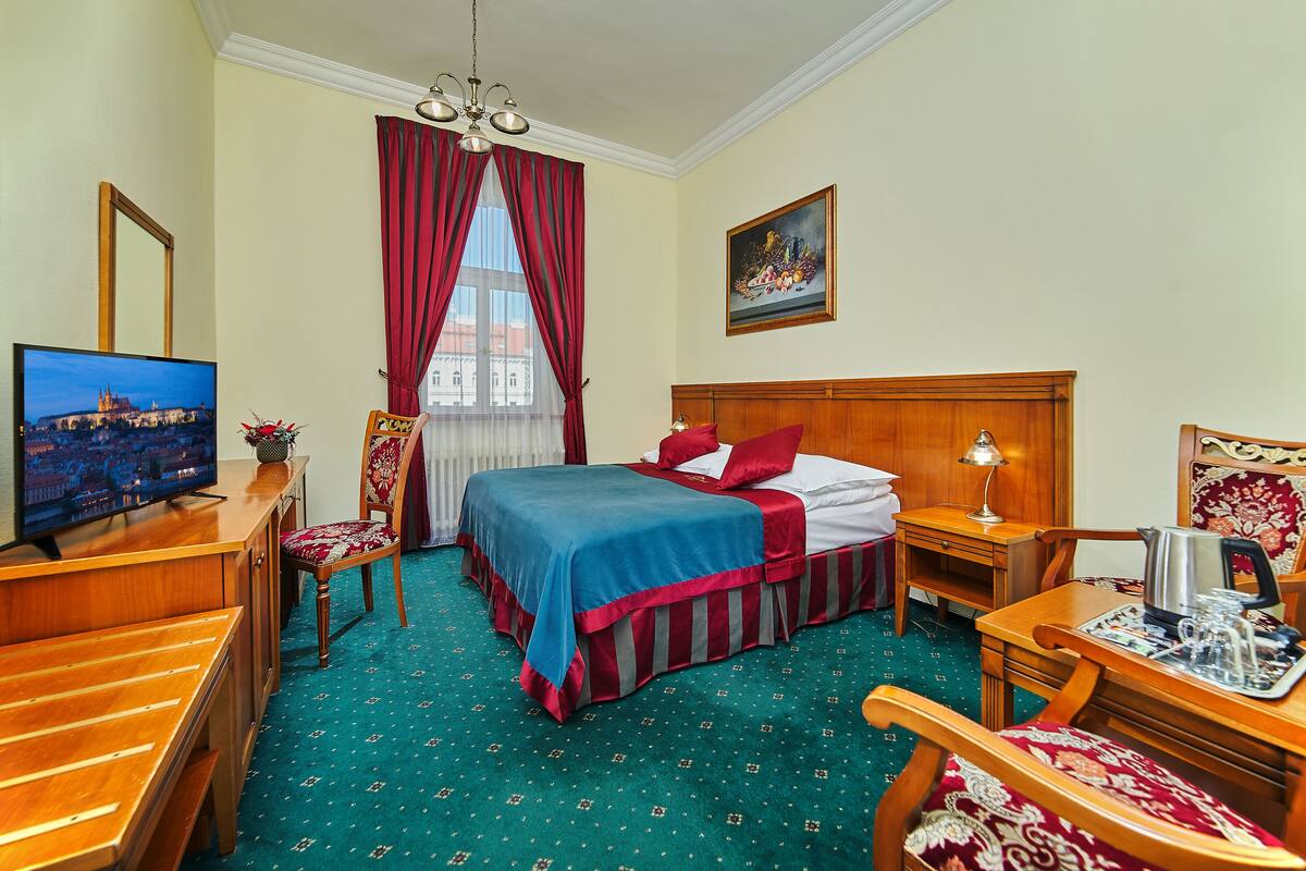 Habitación de hotel con cama, escritorio con televisor, silla, mesa con lámpara y un cuadro en la pared.
