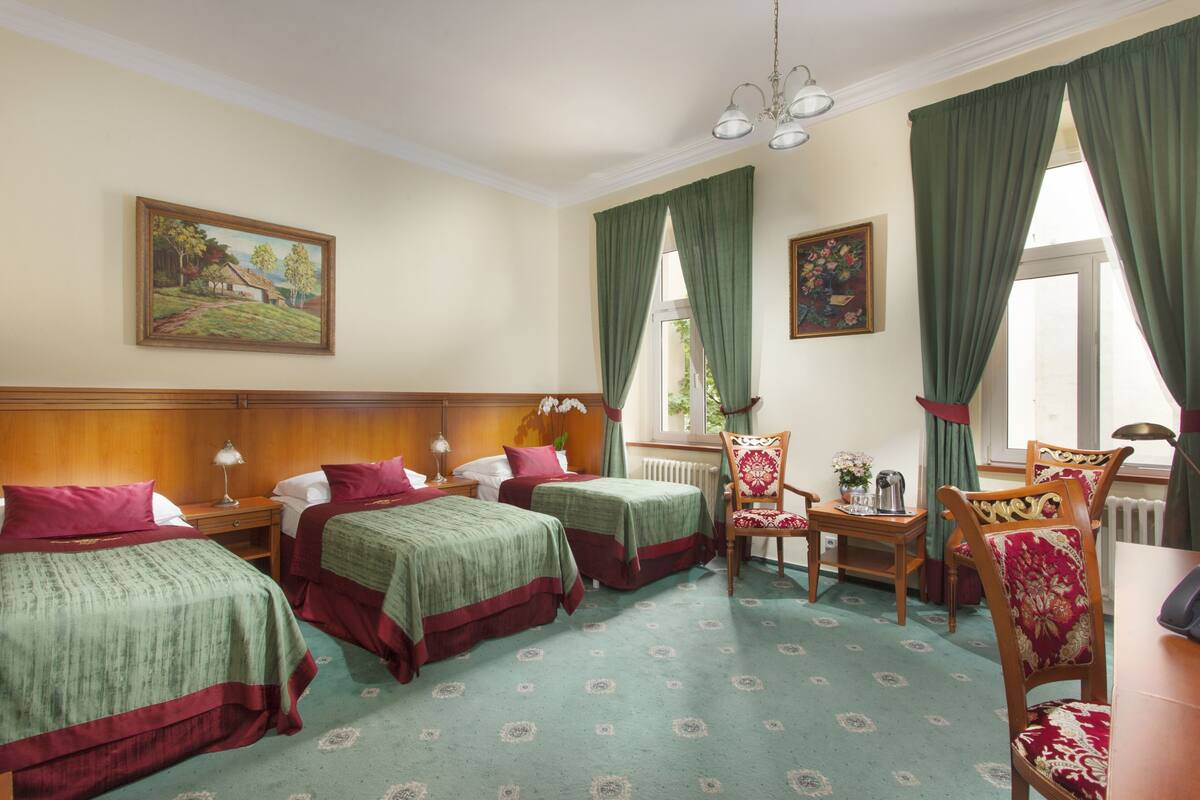 Habitación de hotel con dos camas individuales, cabecero de madera, mesa de comedor con sillas y una silla con cojín estampado de flores.