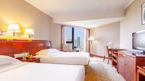 A hotel room with two beds, a desk, a chair, a TV, and a large window with a city view.
