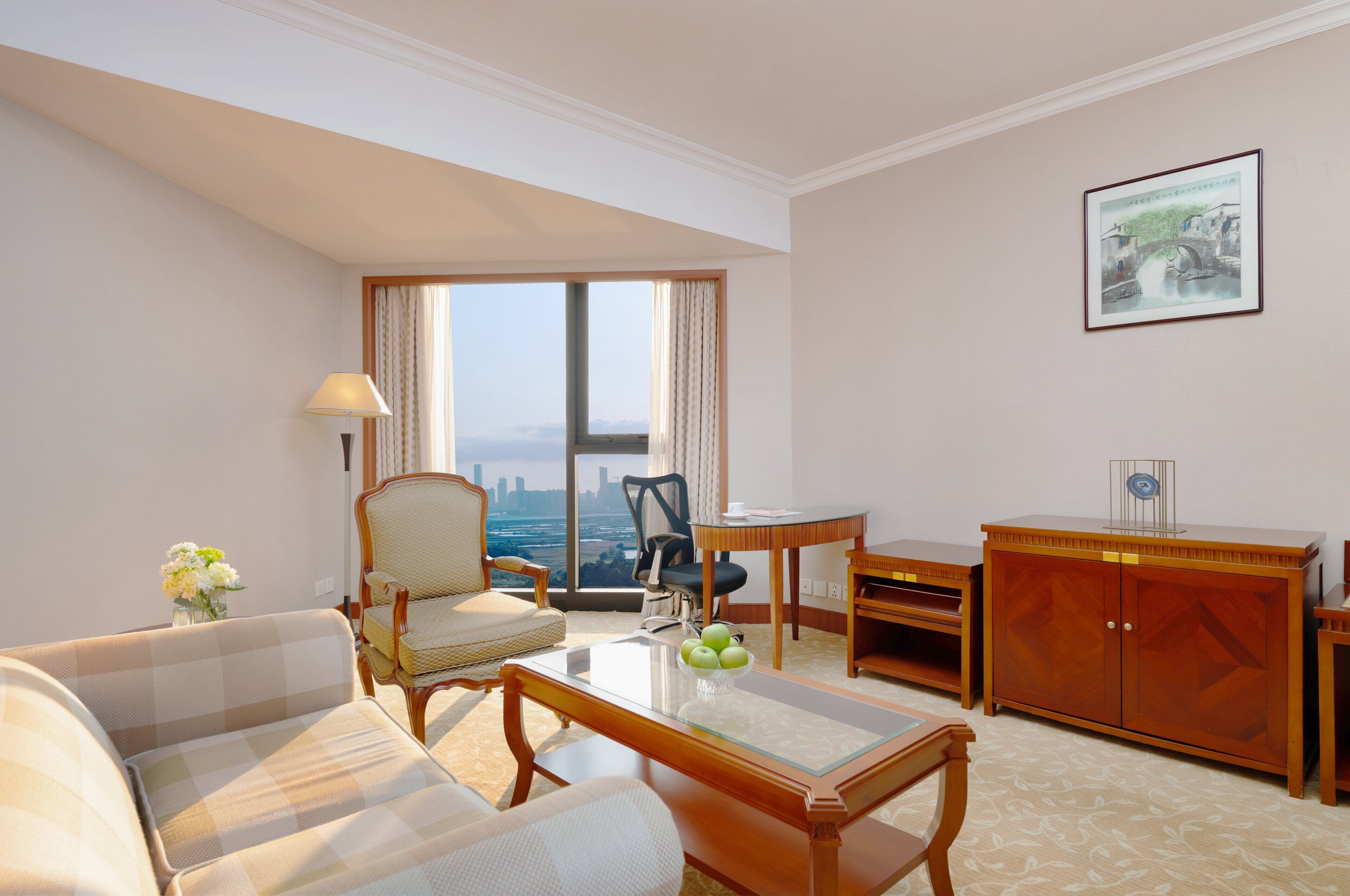 A hotel room with a sofa, armchair, dining table, and a view of a cityscape through large windows.