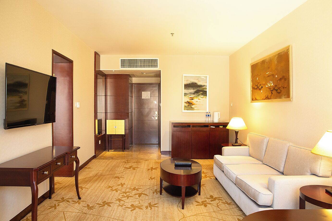 Habitación de hotel con televisión de pantalla plana, sofá, mesa de centro, escritorio, lámpara y obras de arte en las paredes.