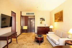 A hotel room with a flat-screen TV, a sofa, a coffee table, a desk, a lamp, and artwork on the walls.