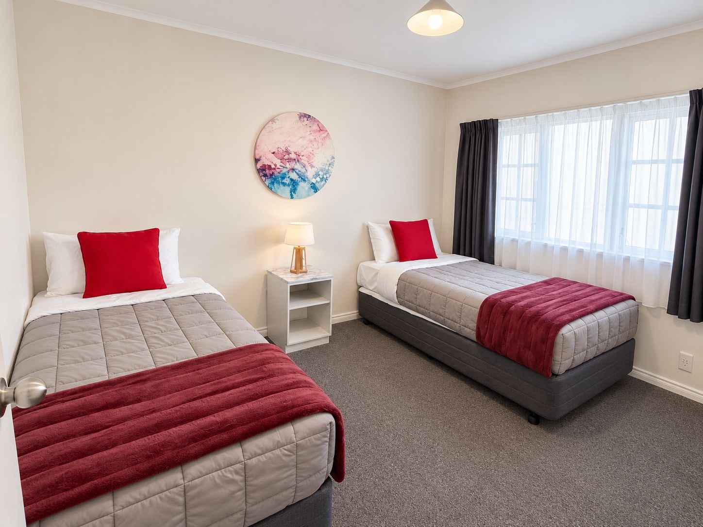 A hotel room with two beds, a nightstand with a lamp, a painting on the wall, and a window with curtains.