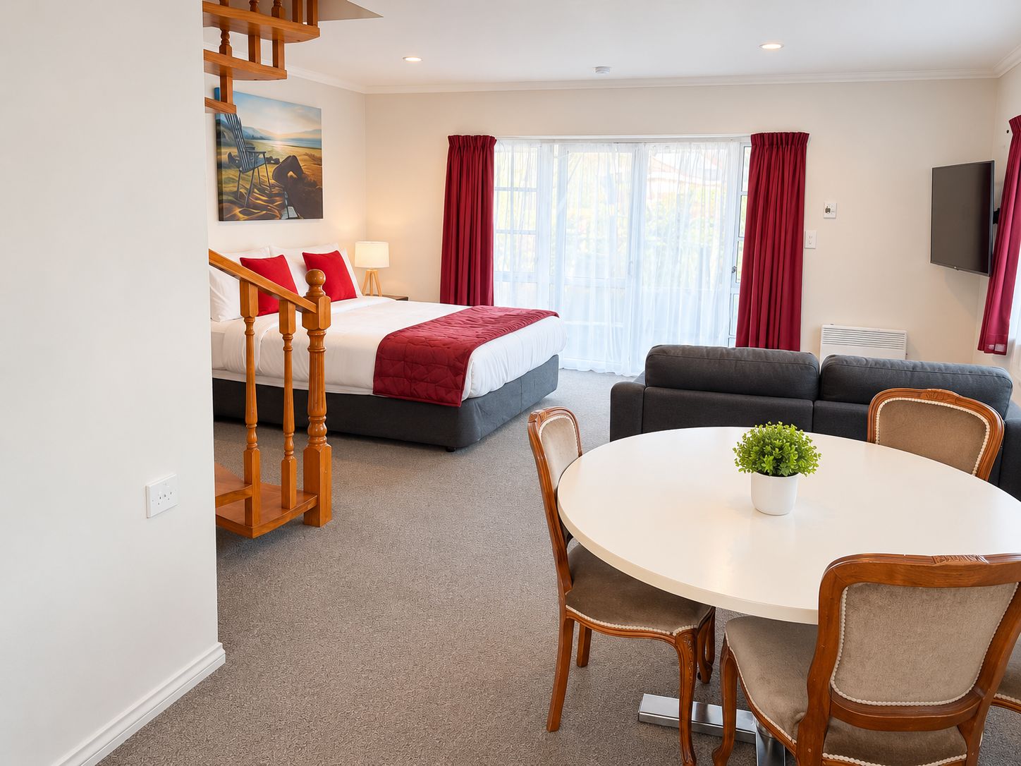 A hotel room with a bed, a sofa, a dining table with chairs, a staircase, and a painting on the wall.