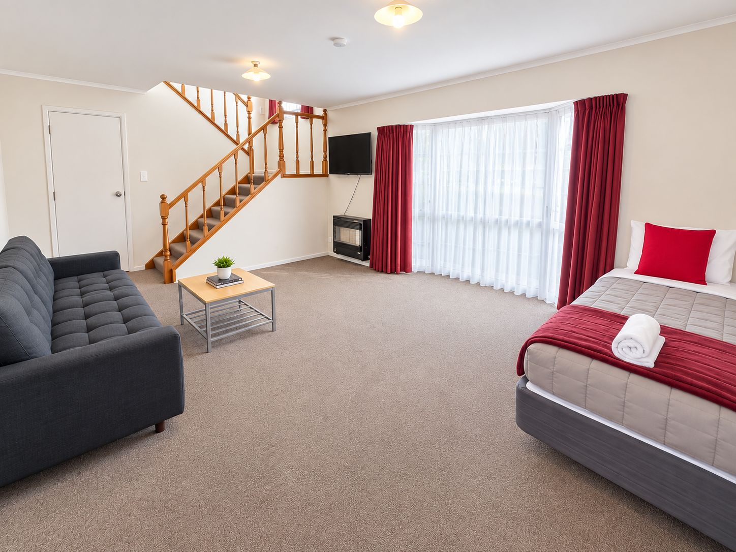 A hotel room with a sofa, a bed with a red blanket, a wooden staircase, a flat-screen TV, and a fireplace.