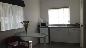 A small, well-lit kitchen with a microwave, a sink, and cabinets. There is a window with sheer curtains and a vase with flowers on the windowsill. - Settlers Motor Lodge (Lower Hutt)